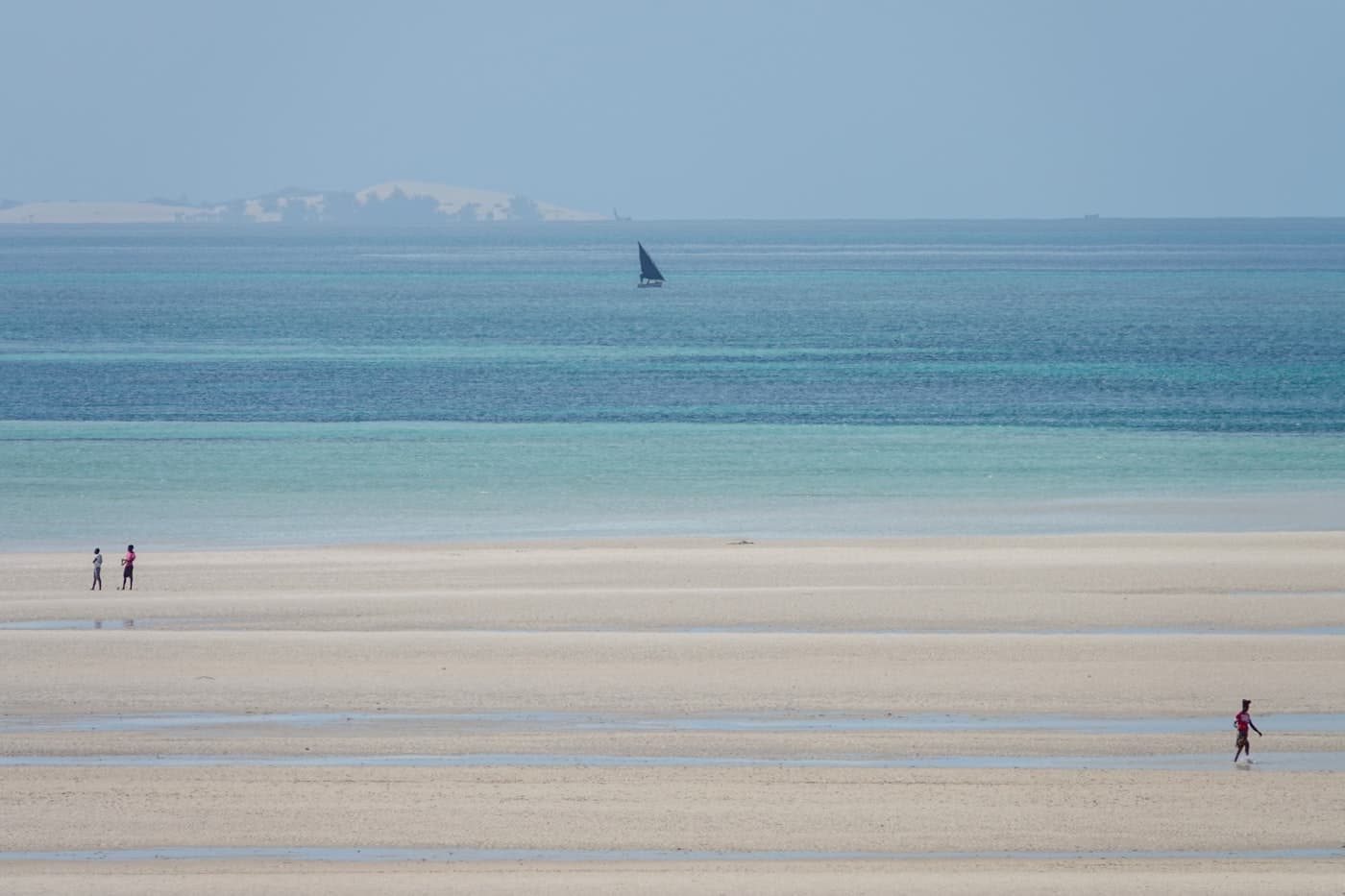 People walk on a sandy beach with a distant sailboat