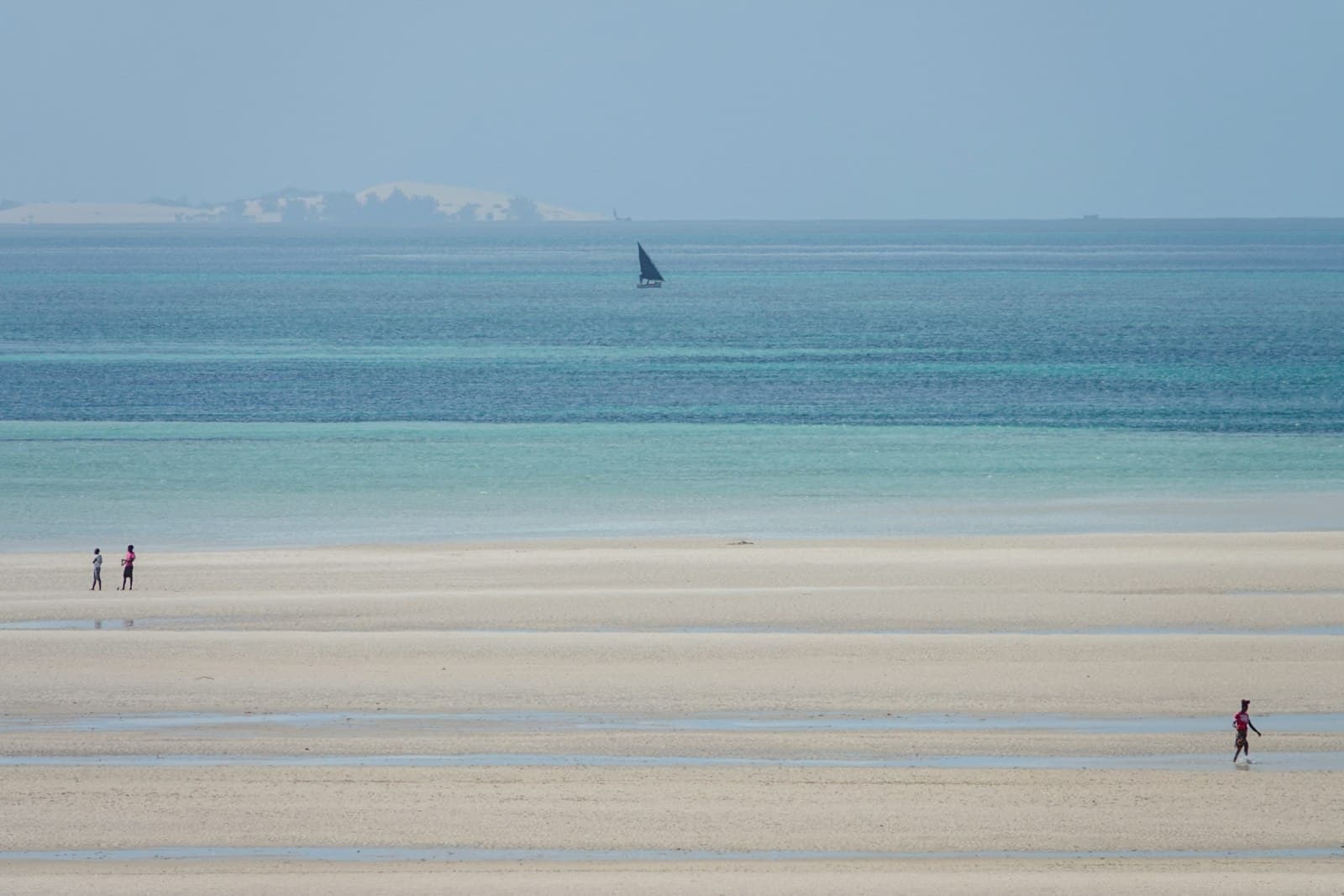 People walk on a sandy beach with a distant sailboat